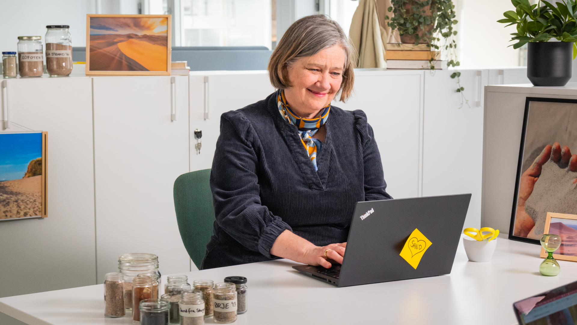 En smilende dame sitter i et kontorlandskap foran en PC og ser ned på skjermen